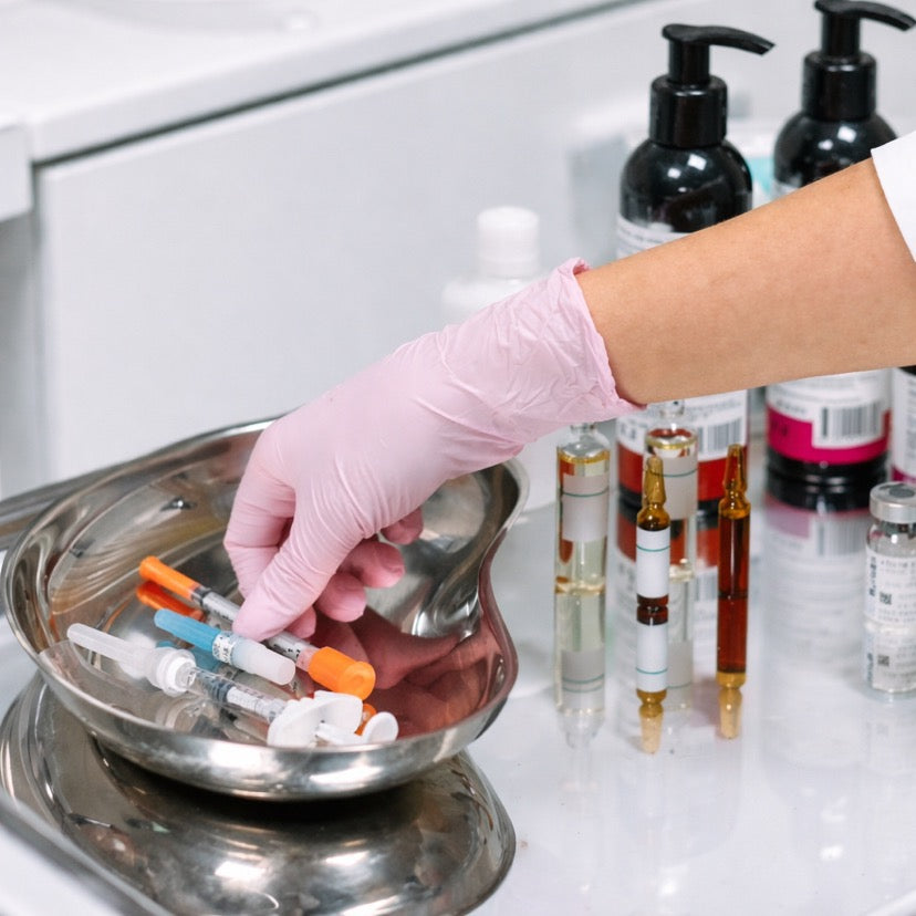 Gloved hand placing cosmetic syringes in a metal tray with skincare vials and bottles